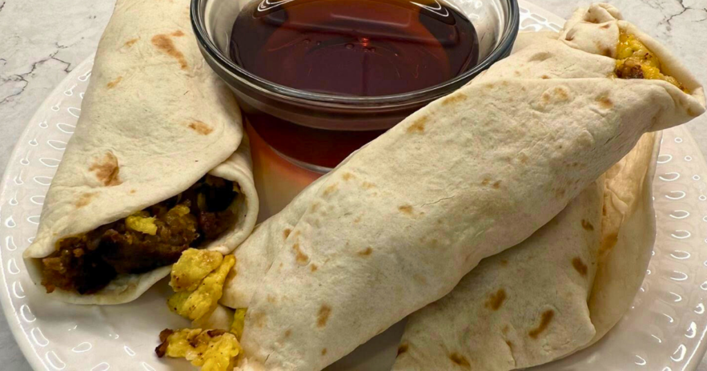Homemade Sausage, Egg, and Cheese Breakfast Burritos sitting on a white plate with a cup of syrup.